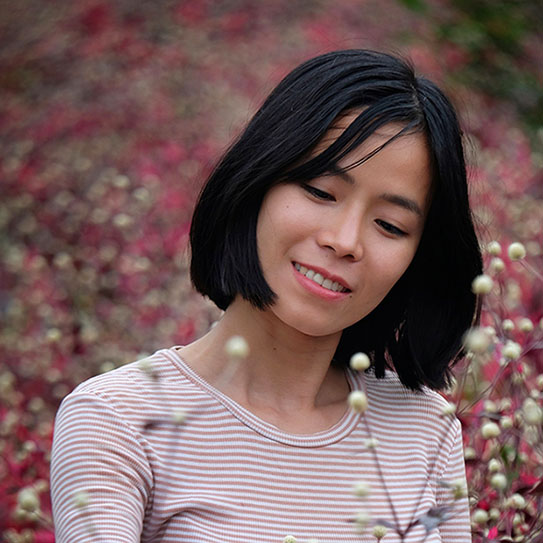 A woman with short black hair and a striped shirt stands outdoors, surrounded by pink and white flowers, looking down and smiling softly—much like the friendly atmosphere you’ll find at Newton MA dentist offices.
