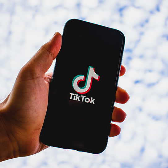 A hand holds a smartphone displaying the TikTok app logo on the screen against a blurred, light-colored background, perfect for newton ma dentist offices or dentists near Boston looking to connect with patients through social media.