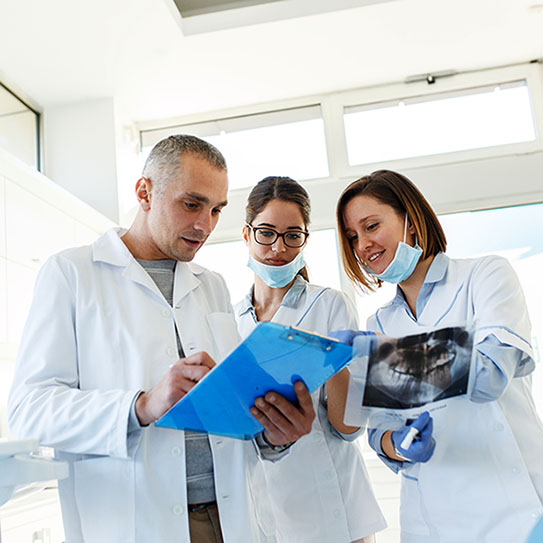 Three medical professionals in lab coats and masks review a dental X-ray and discuss notes on a blue clipboard in a bright examination room, exemplifying the expertise of Newton dentists near Boston.