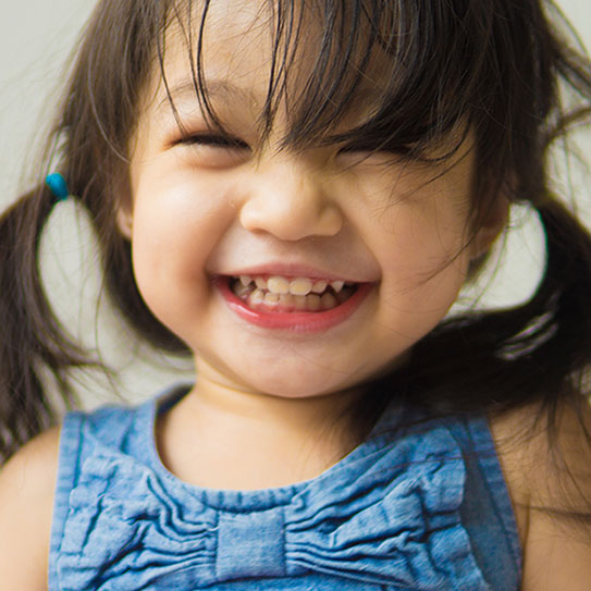A young child with pigtails and a blue top smiles widely, showing teeth, with hair partially covering their face—radiating the joy seen after a visit to Newton dentists near Boston.